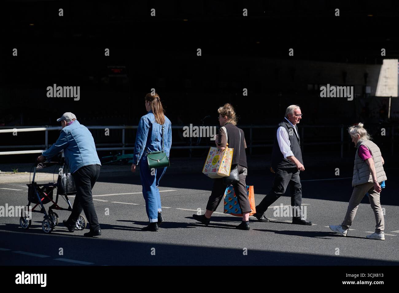 Seitenansicht einer Gruppe gemischter Generationen, die die Stadtstraße überquert, ältere Menschen mit Gehhilfe, helles Sonnenlicht, horizontaler Rahmen, Editorial für demografische Daten, Urban in Stockfoto