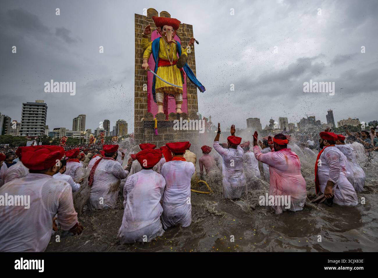 Mumbai, Indien - 6. September 2025: Blick auf eine große Statue von Ganesha, inmitten jubelnder Gläubiger, die durch das Wasser waten, deren rote Hüte sich vom bewölkten Himmel abheben. Stockfoto