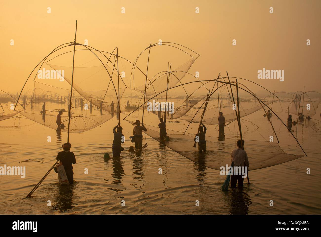 Polo, Bangladesch - 28. November 2023: Blick auf Figuren, die durch seichtes Wasser waten, die Landschaft von Ruhul Bill mit komplizierten Fischereistrukturen unter einem trüben, goldenen Himmel. Stockfoto