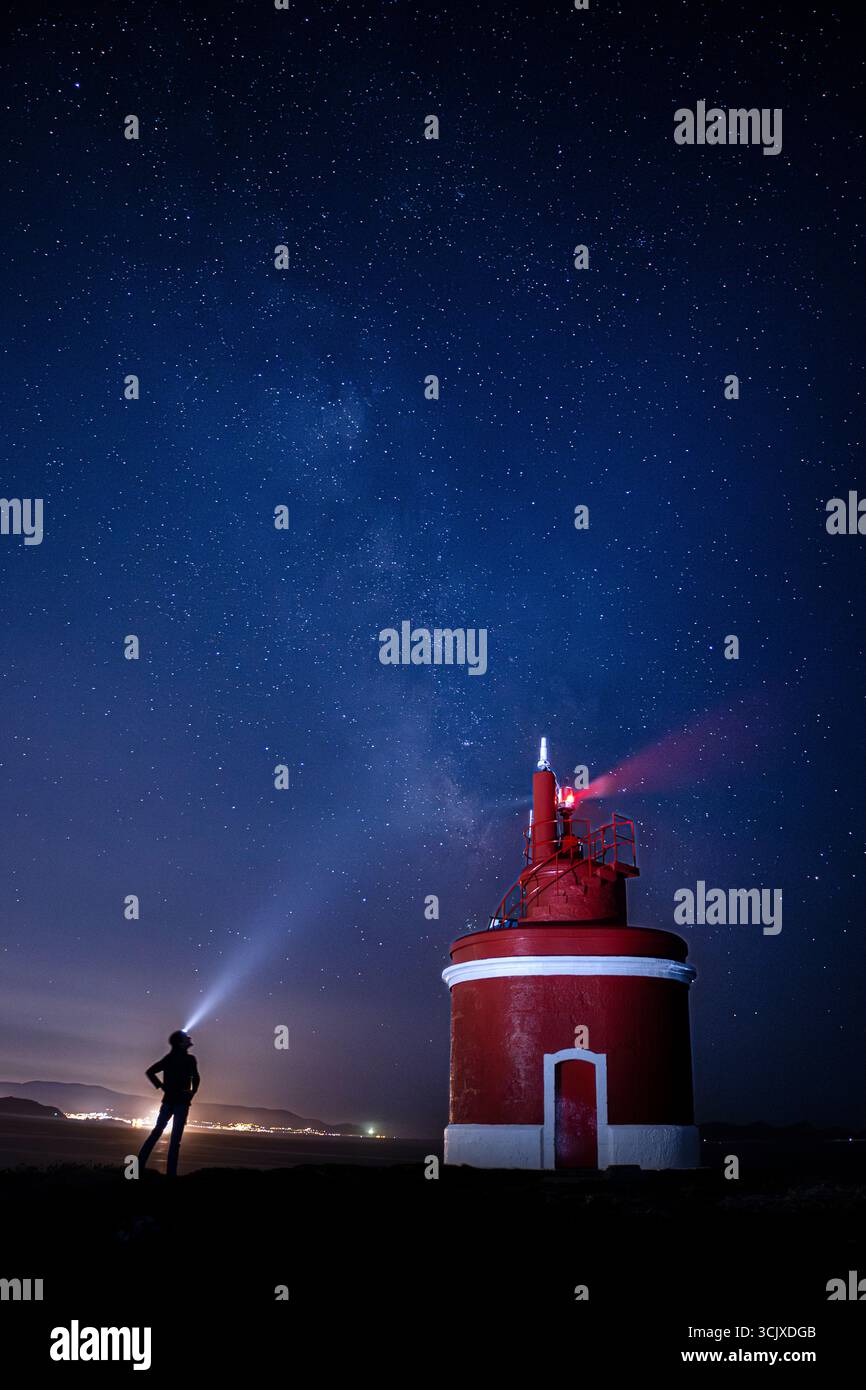 Unter einem riesigen, sternenübersäten Nachthimmel steht eine Person in Silhouetten und strahlt einen hellen Lichtstrahl von einem Scheinwerfer in Richtung Himmel. Zu ihrer Rechten, Stockfoto
