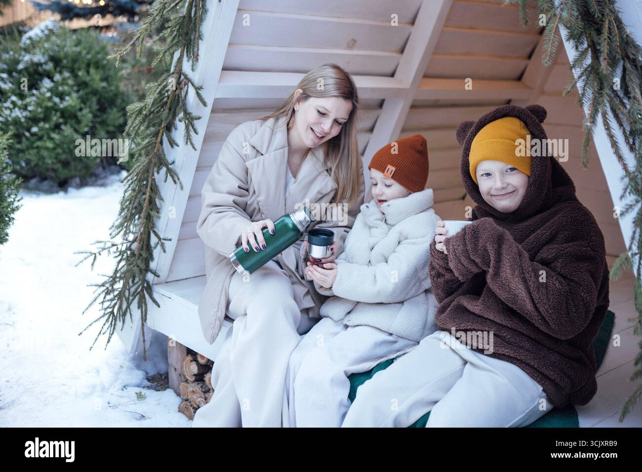 Mutter und zwei Kinder teilen sich warme Getränke in Winterkleidung, eingebettet in einen bezaubernden Außenbereich, mit Schnee und Grün, die das gemütliche Ambiente unterstreichen Stockfoto