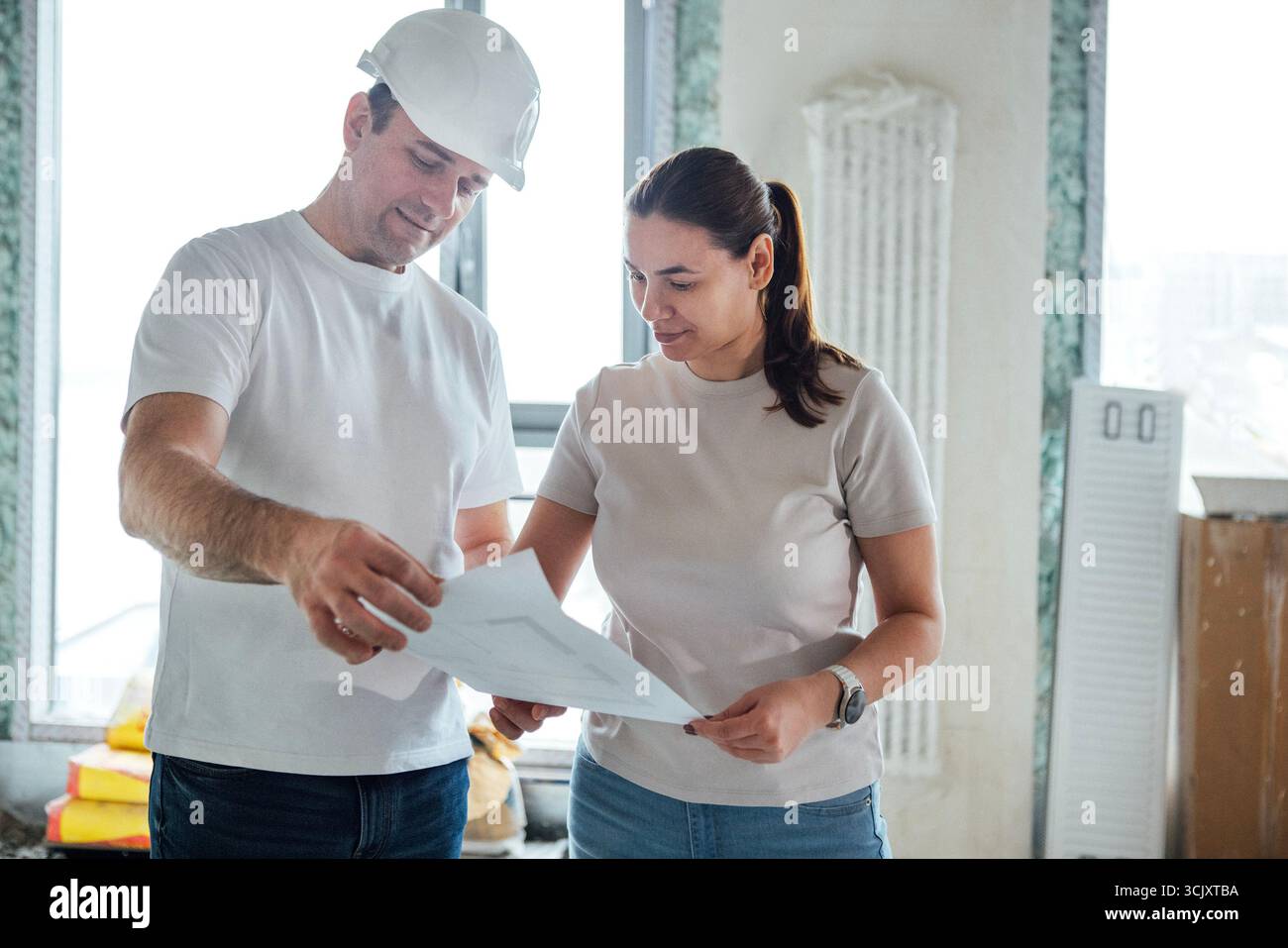 Mann und Frau prüfen Baupläne in einem gut beleuchteten, unvollendeten Raum mit Werkzeugen und Materialien, wobei Teamarbeit und Projektplanung hervorgehoben werden Stockfoto