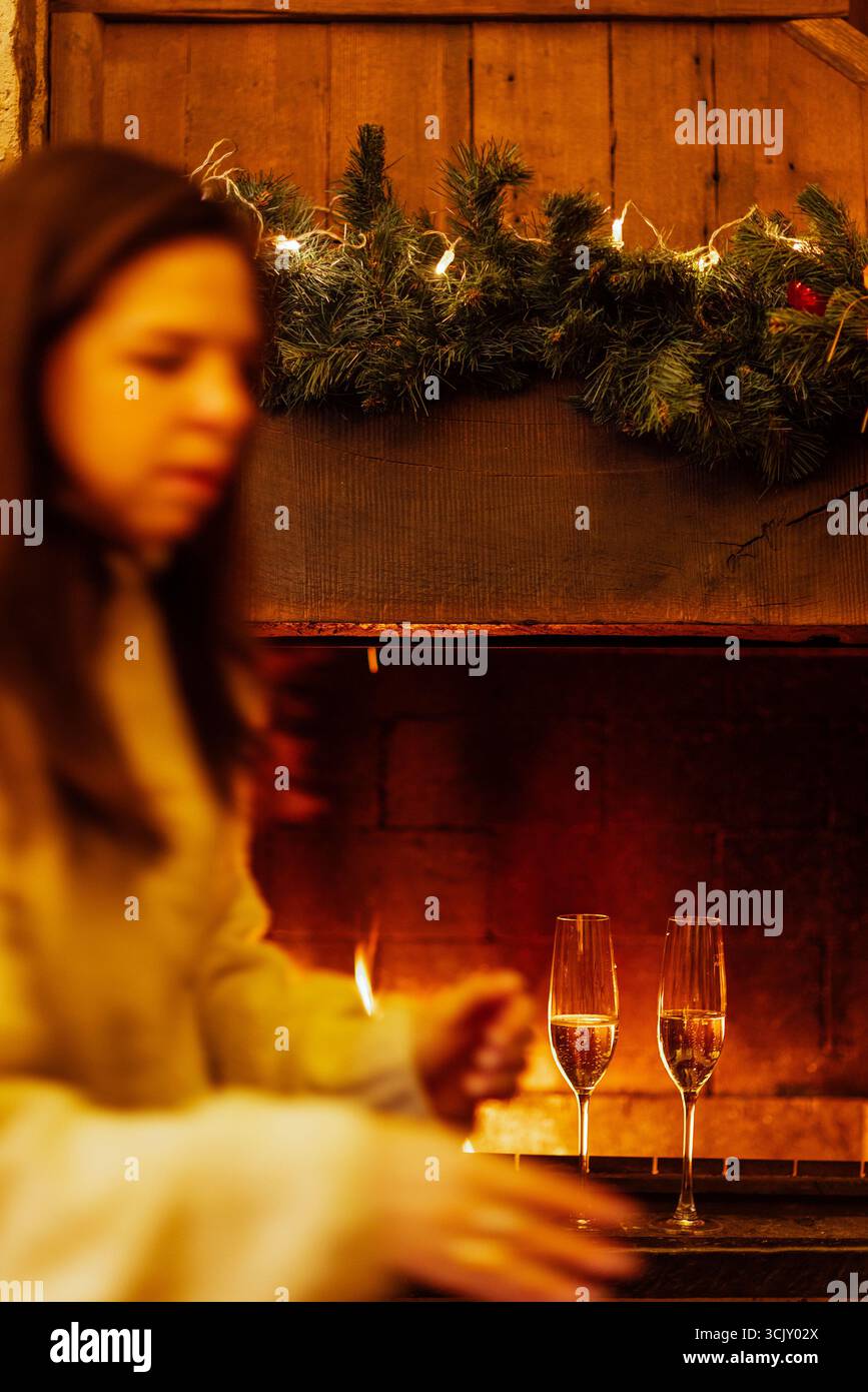 Frau in weichem Pullover bereitet sich auf eine festliche Feier vor einem warmen Kamin, dekoriert mit Grün und Lichtern, die ein gemütliches Urlaubsambiente hervorrufen Stockfoto