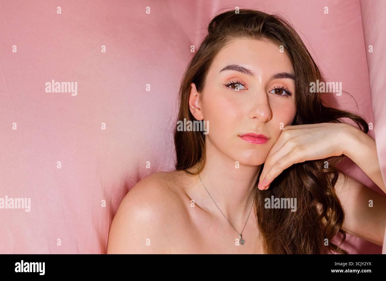 Weibliches Model mit langen Haaren posiert vor zartem rosa Hintergrund und unterstreicht ihre natürliche Schönheit und ihr ruhiges Auftreten in einer ruhigen und einladenden Atmosphäre Stockfoto