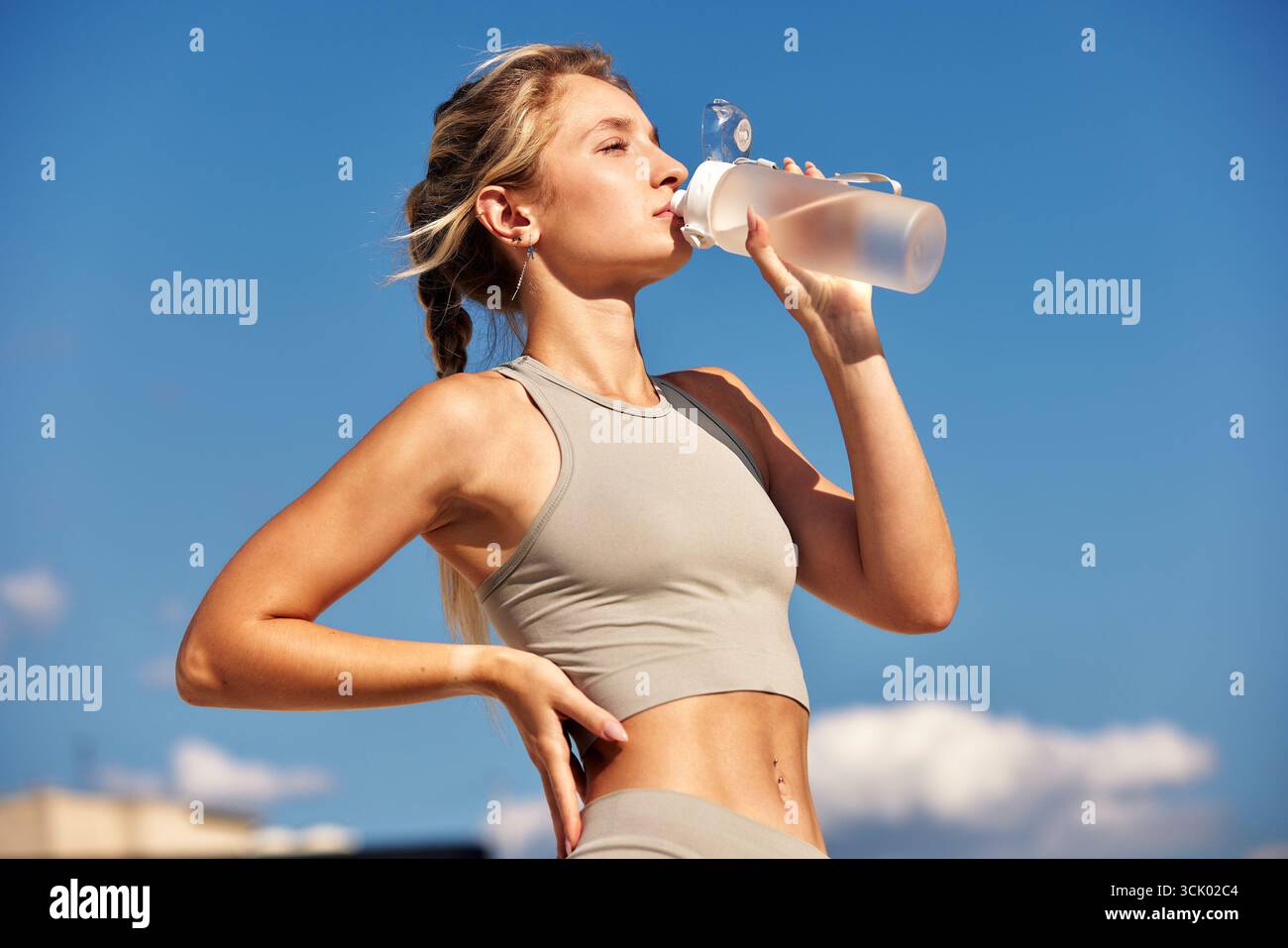 Eine fitte junge Frau genießt ein erfrischendes Getränk aus einer Flasche im Freien unter einem klaren blauen Himmel. Sie trägt sportliche Kleidung und scheint zufrieden zu sein Stockfoto