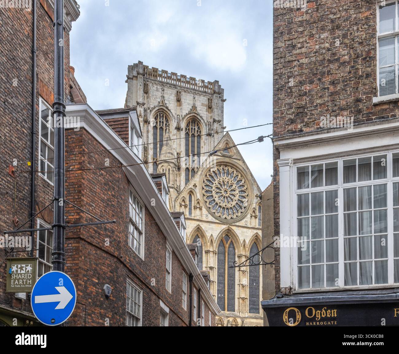 Blick auf York Minster von einer engen Straße in York, England, mit Backsteinbauten, Ladenschildern und gotischer Architektur im Hintergrund Stockfoto