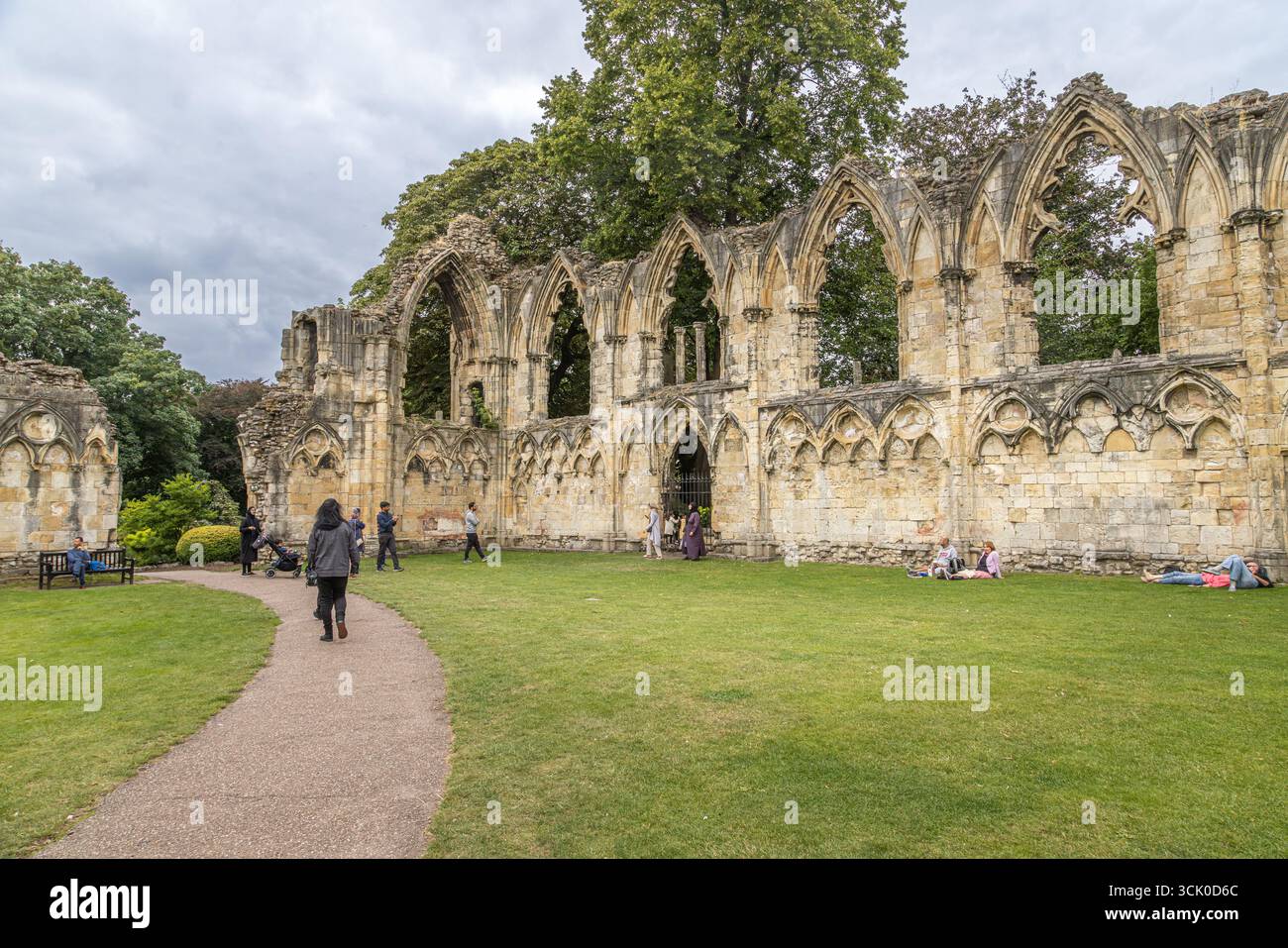 Ruinen der St Mary’s Abbey in York Museum Gardens, England, mit gotischen Bögen, verwittertem Steinwerk, grasbewachsenen Rasenflächen, und Menschen, die laufen. Stockfoto