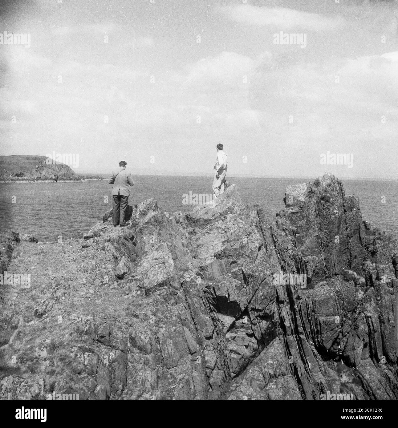 1960er Jahre, historisch, zwei Erwachsene Männer stehen auf zerklüfteten Felsen an der Küste und blicken auf den Ozean. Stockfoto