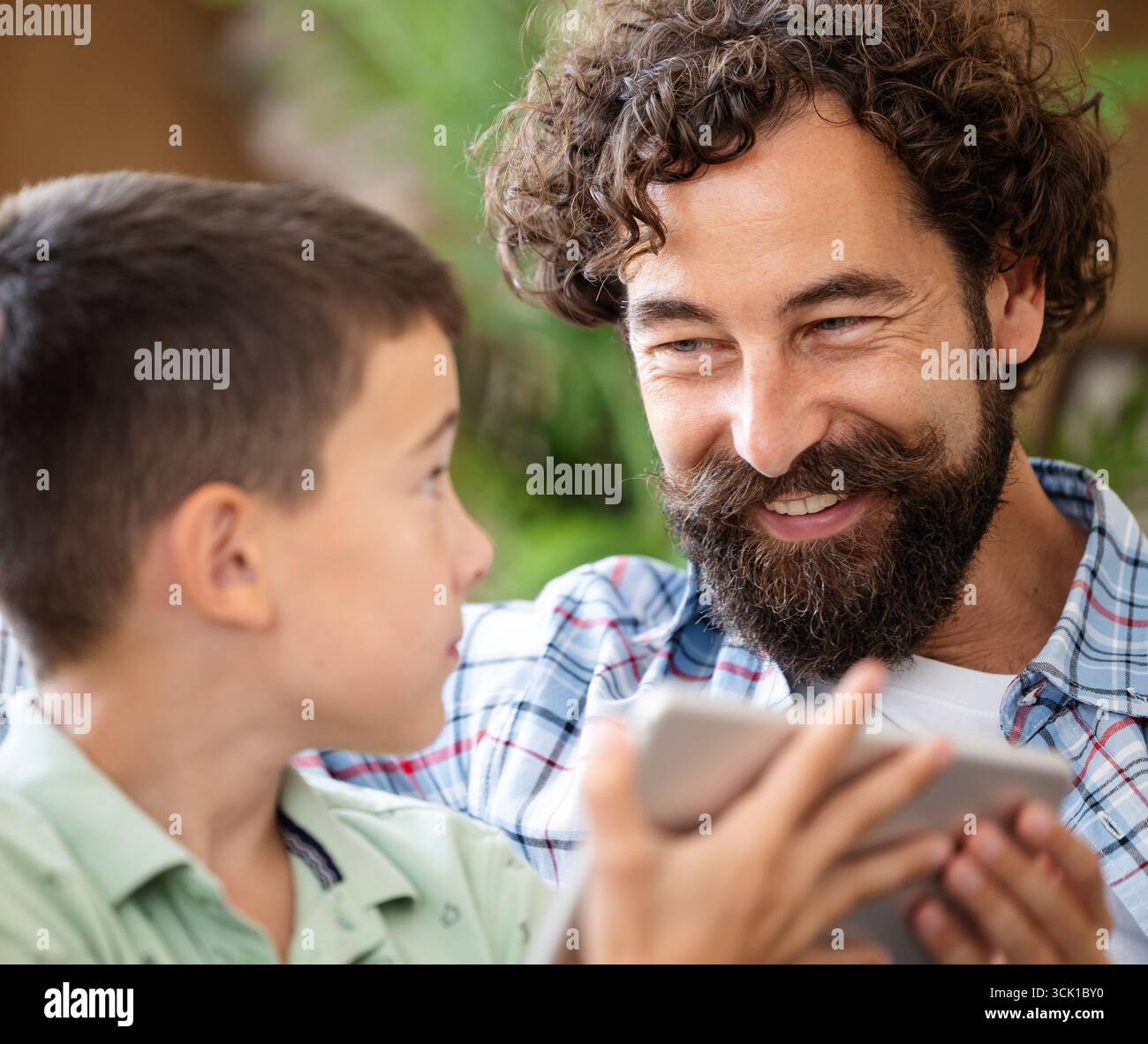 Vater und Sohn teilen freudige Momente, während sie mit dem Tablet in entspannter, natürlicher Umgebung interagieren. Sie lächeln einander an Stockfoto