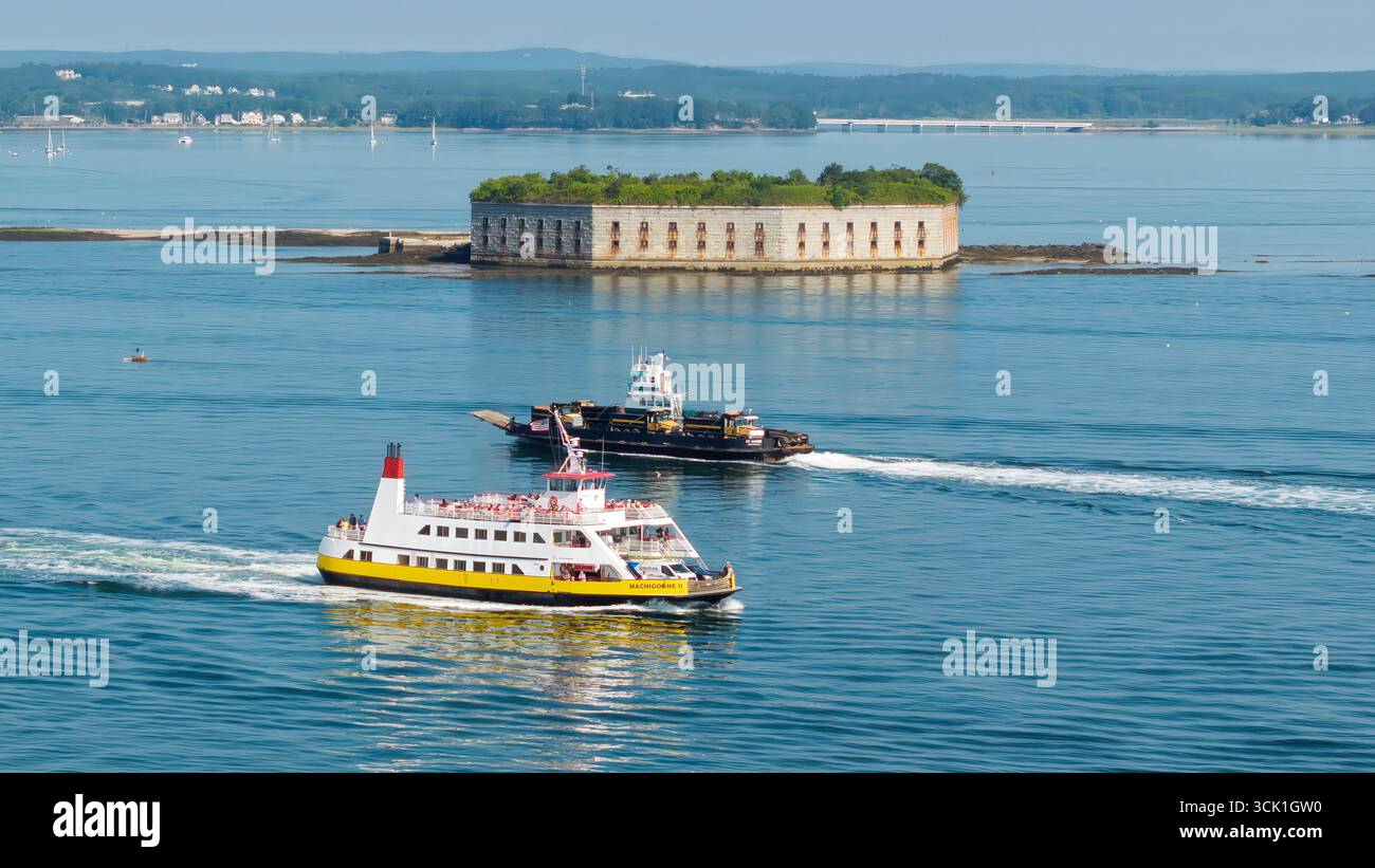 Aus der Vogelperspektive einer leuchtend gelb-weißen Fähre, die durch das ruhige Wasser von Portland, Maine, gleitet und einen weichen Weg hinter sich lässt. In der Ferne, ein streng Stockfoto