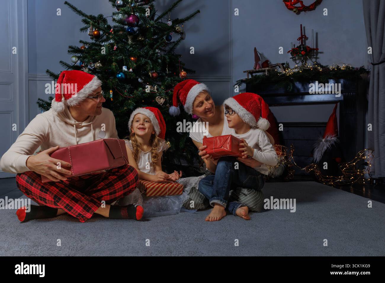 Familie in Ferienhüten sitzt neben dem Baum, hält gewickelte Geschenke Stockfoto