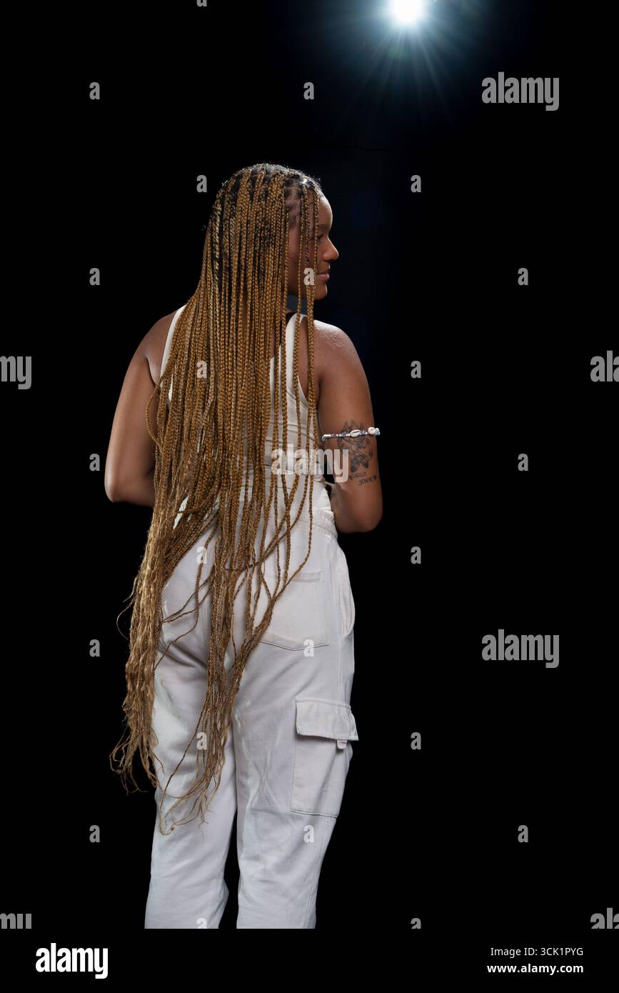 Salvador, Bahia, Brasilien - 30. August 2025: Junge Frau mit geflochtenem Haar, die mit dem Rücken zur Kamera steht und für ein Studiofoto gegen einen Blac posiert Stockfoto