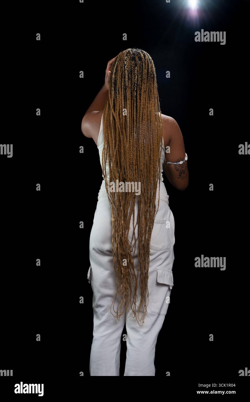 Salvador, Bahia, Brasilien - 30. August 2025: Junge Frau mit geflochtenem Haar, die mit dem Rücken zur Kamera steht und für ein Studiofoto gegen einen Blac posiert Stockfoto