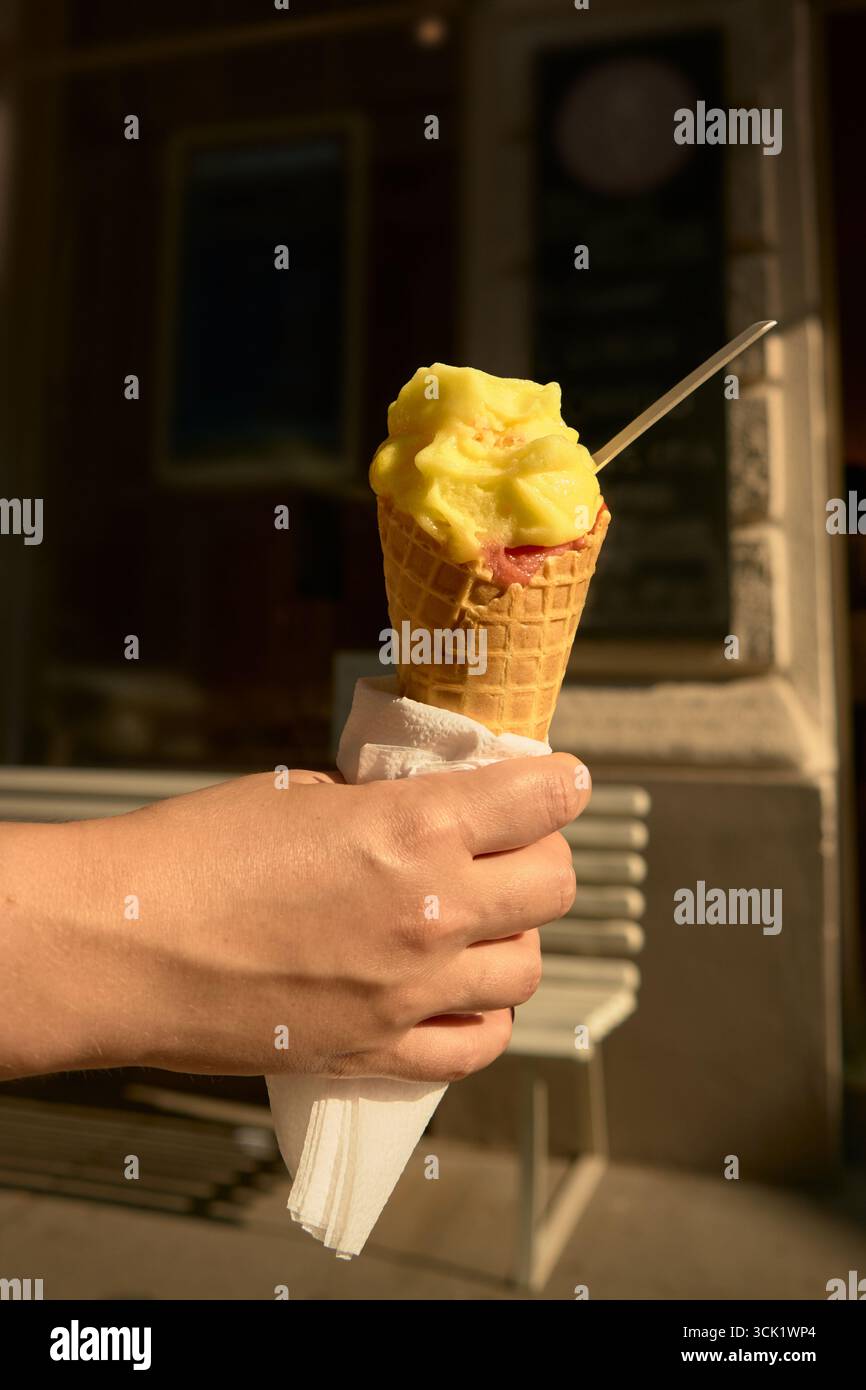 Hand hält gelbe und rosafarbene Eiskegel an einem sonnigen Tag, mit Holzbank im Hintergrund, symbolisiert Sommer-Vibes, süße Leckereien und relaxa im Freien Stockfoto