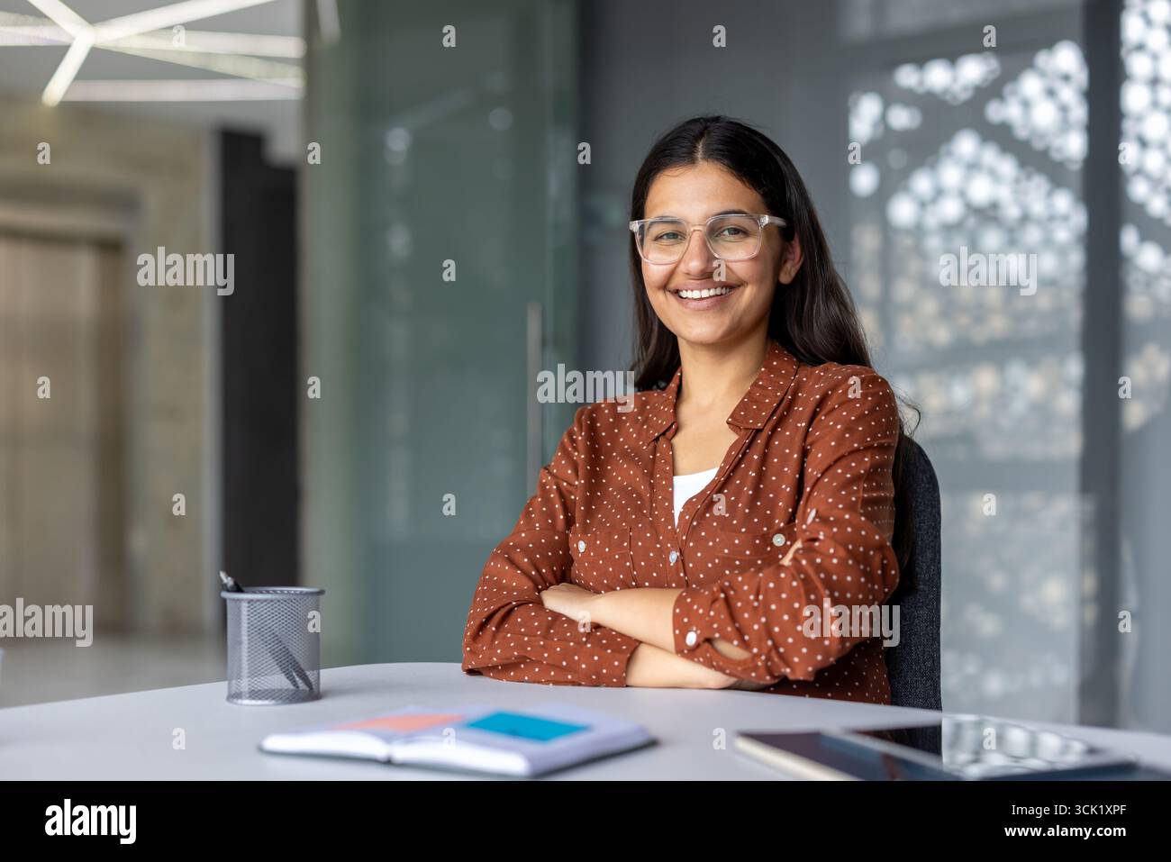 Die junge indische Geschäftsfrau lächelt selbstbewusst mit gekreuzten Armen, während sie an einem modernen Schreibtisch sitzt, und strahlt Professionalität und Ehrgeiz in einer stilvollen Unternehmensumgebung aus Stockfoto