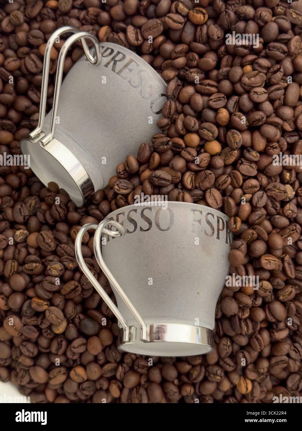 Eine Hochwinkelaufnahme von zwei Espresso-Tassen, die auf einem Bett mit gerösteten Kaffeebohnen liegen. Das Bild hebt die Textur und die tiefbraune Farbe der Bohnen hervor, weiß - Smartphone-aufgenommenes Stockfoto