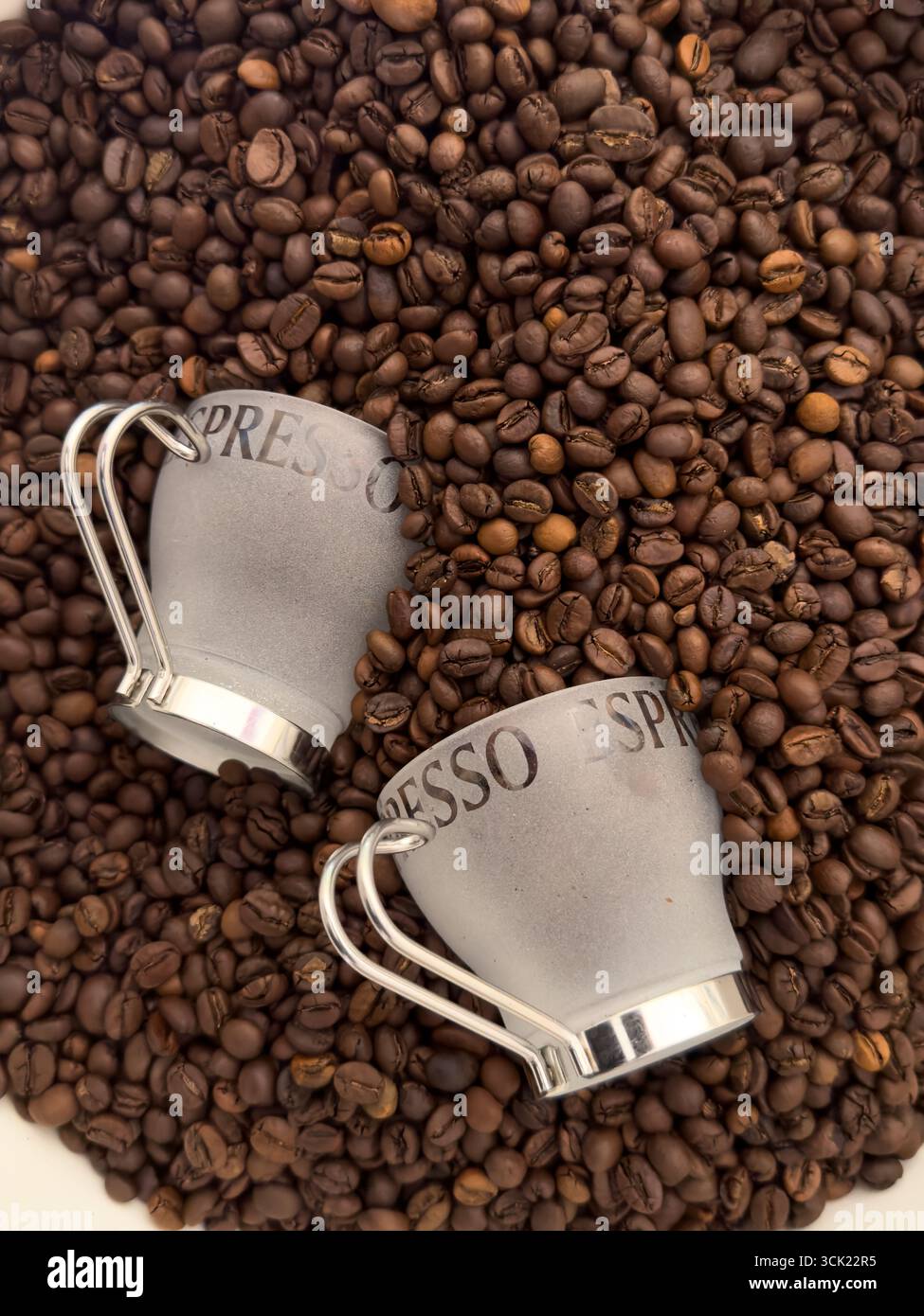 Eine Hochwinkelaufnahme von zwei Espresso-Tassen, die auf einem Bett mit gerösteten Kaffeebohnen liegen. Das Bild hebt die Textur und die tiefbraune Farbe der Bohnen hervor, weiß - Smartphone-aufgenommenes Stockfoto