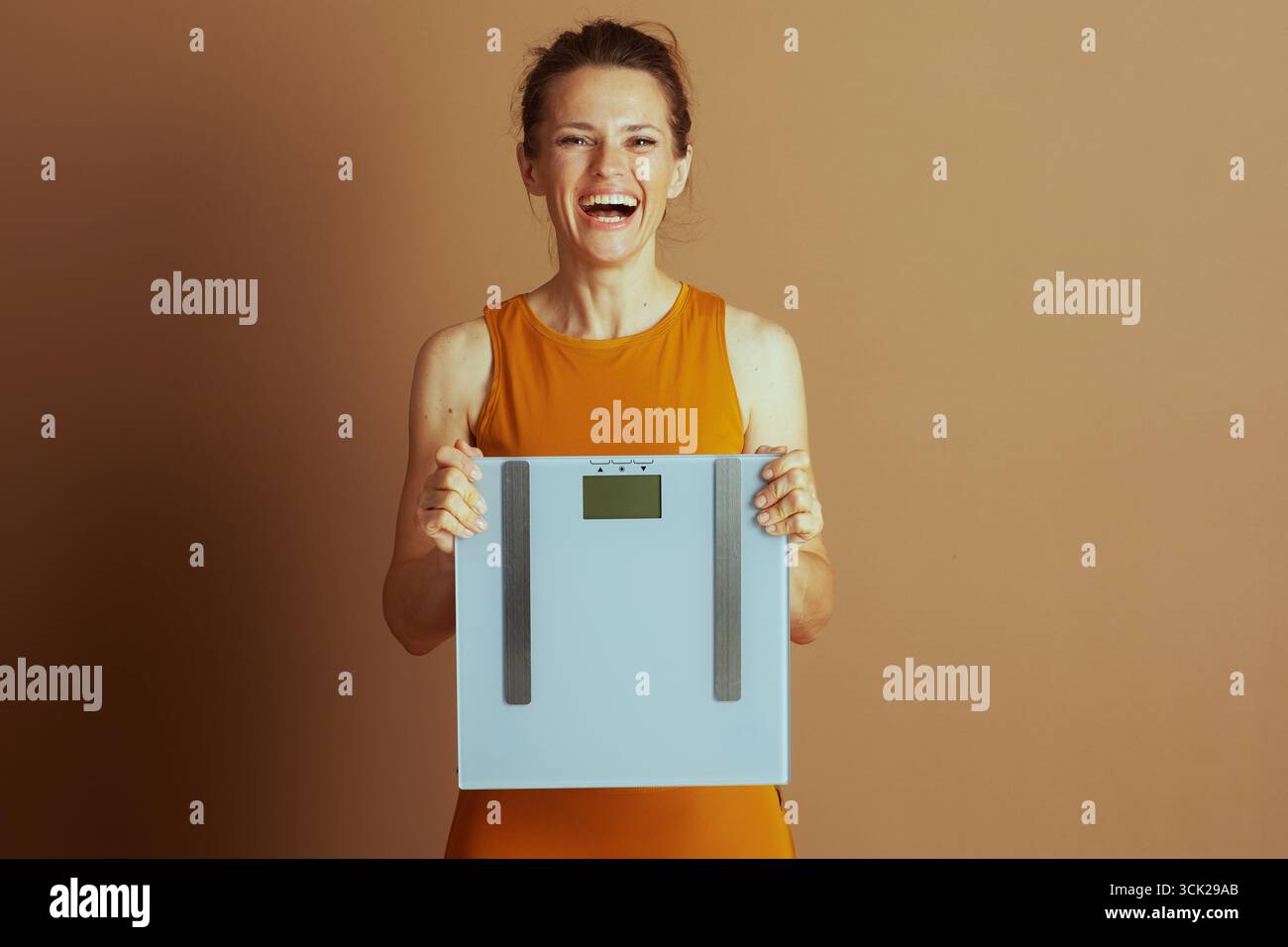 Eine freudige Frau in einem orangen Tanktop lacht, hält eine digitale Skala, symbolisiert Glück, Leistung und positive Ergebnisse in ihrer Gesundheit, Fitness, Stockfoto