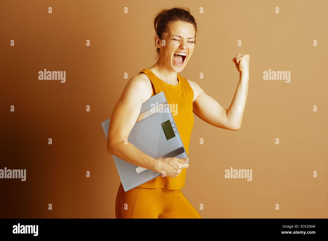 Die Frau in orangefarbenem Sportoutfit schreit aufgeregt, pumpt triumphierend die Faust, während sie die digitale Waage hält. Dieses Bild vermittelt überwältigende Freude Stockfoto