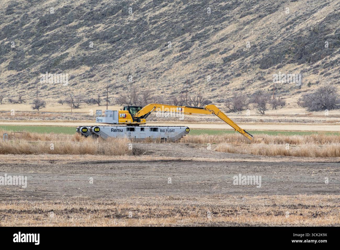 Tulelake, CA/USA – 29. Dezember 2023: Ein Caterpillar-Bagger für schwere Maschinen auf dem amphibischen Remu Big Float (E22) mit Buschland im Hintergrund. Stockfoto