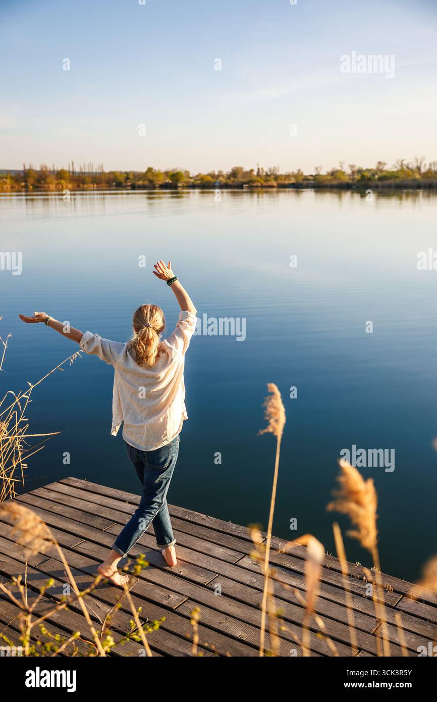 Sorglose Frau, die auf dem Pier am See steht und mit den Armen in fröhlicher Pose tanzt und die ruhige Atmosphäre des indischen Sommers genießt Stockfoto