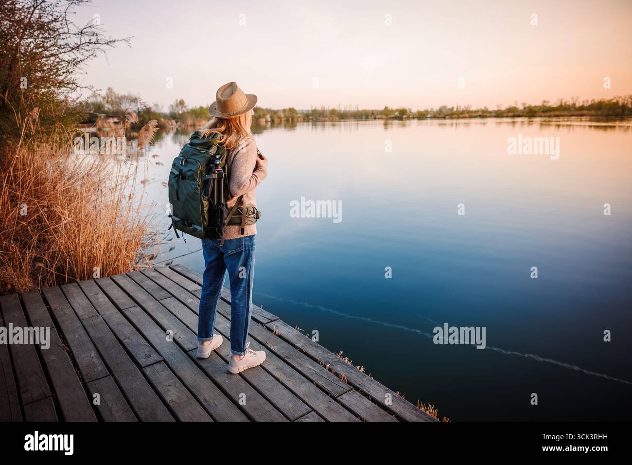 Frau mit Rucksack, die während des friedlichen Sonnenuntergangs im indischen Sommer vom Pier aus auf den ruhigen See blickt. Wanderer, der sich während des Wanderns und des Ökotourismus ausruhen kann Stockfoto