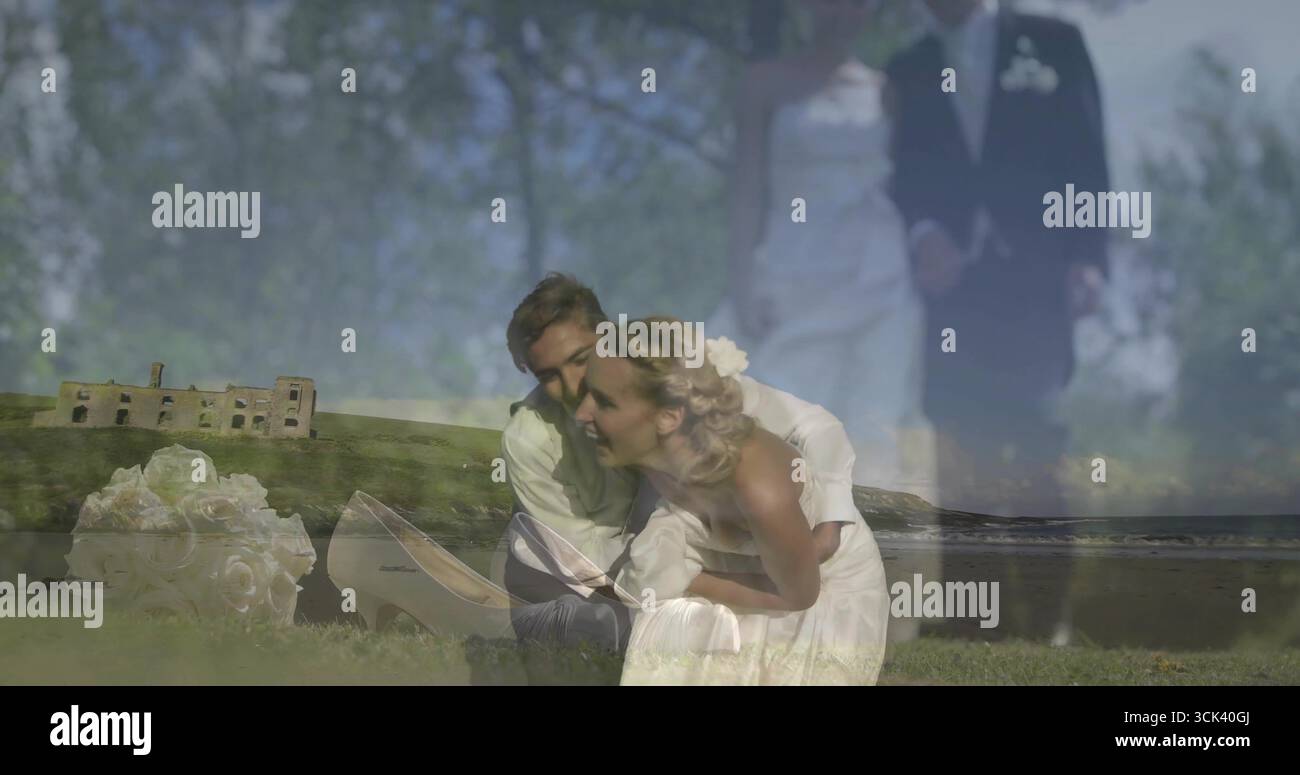 Kniende Braut in weißem Kleid, umarmt mit Bräutigam im Anzug an der grasbewachsenen Küste, mit Blumenstrauß, Schuhen Stockfoto