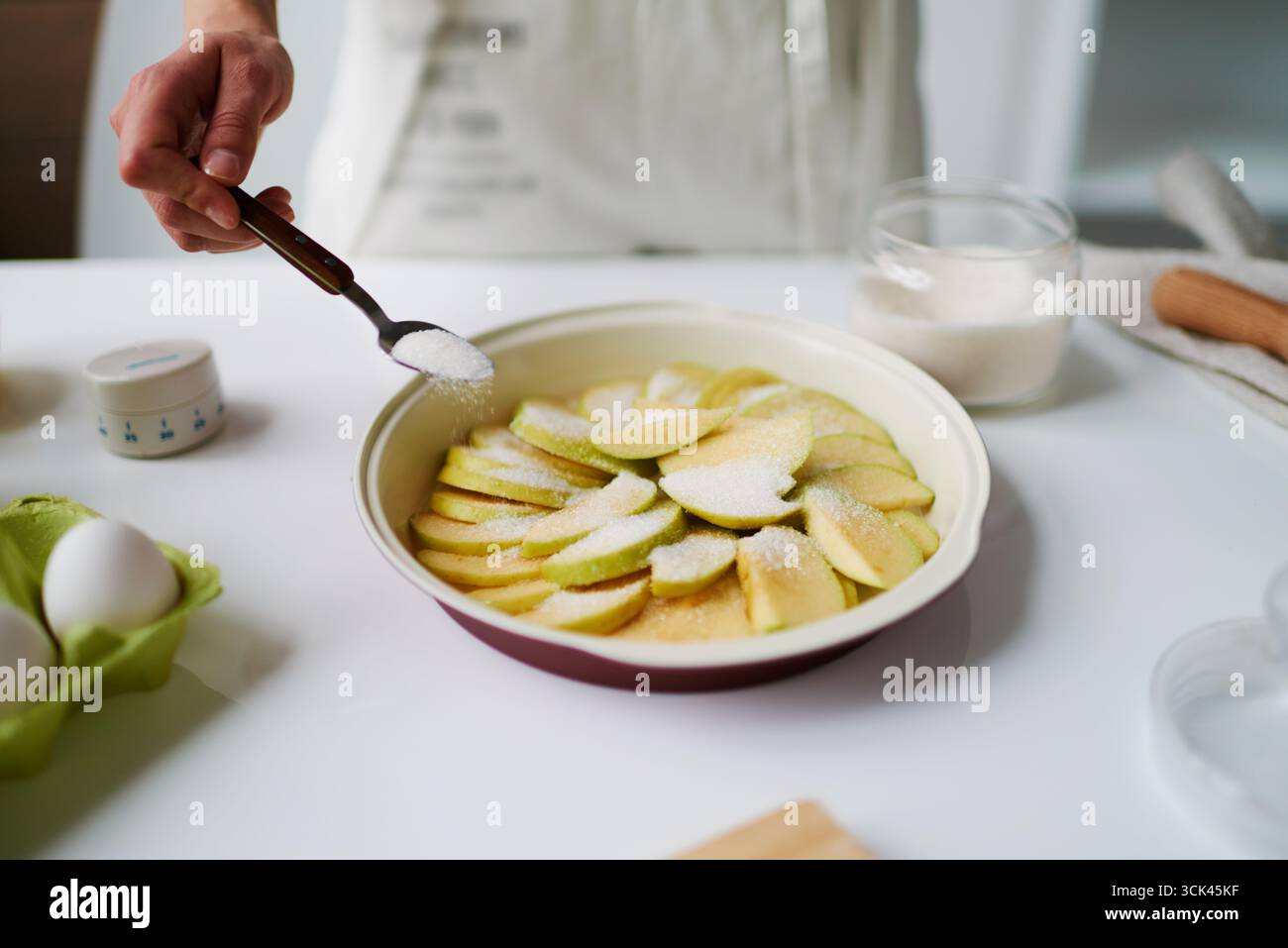 Unbekannte Frau streut Zucker über geschnittene Äpfel in der Backform, bereitet Dessert in der Küche, Eier und Glasschale mit Zucker sichtbar auf der Arbeitsplatte Stockfoto
