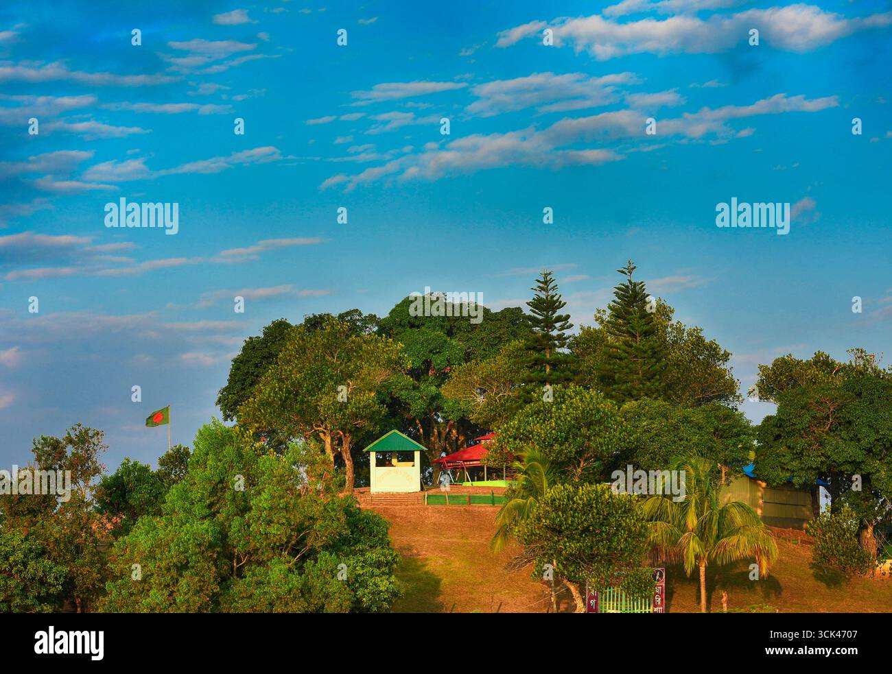 Sajek, Bangladesch - 27. Januar 2019: Blick auf einen üppig grünen Hügel, gekrönt von lebhaften Bäumen, ein kleines weißes Gebäude und die flatternde Flagge Bangladeschs vor einem hellblauen Himmel. Stockfoto