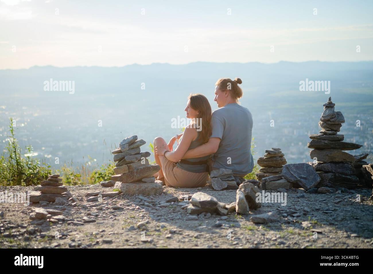 Romantisches Paar, das von hinten umarmt sitzt und den atemberaubenden Blick auf die Stadt vom Gipfel des Berges aus genießt. Perfekt für Reisen, Wandern und Liebesgeschichten. Stockfoto