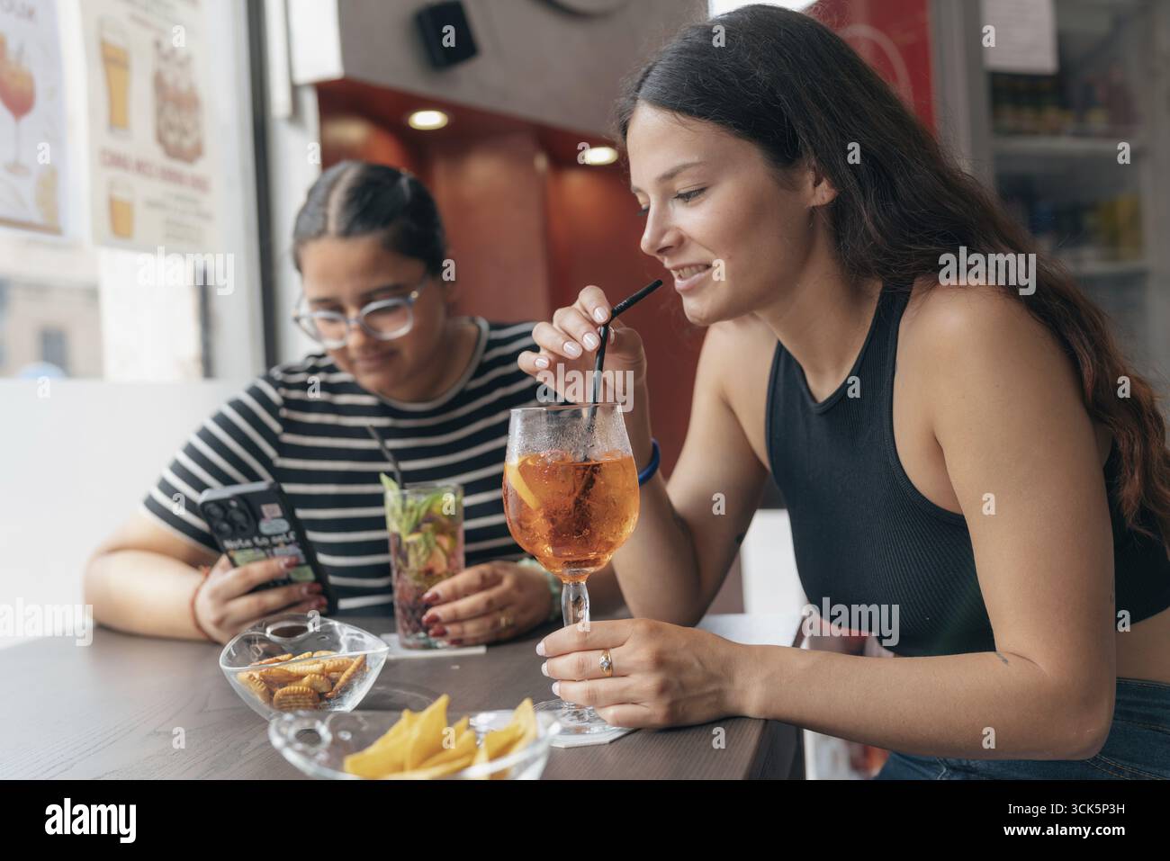 Zwei junge Frauen genießen erfrischende Cocktails und Snacks in einer lebhaften Café-Bar mit zwangloser und geselliger Atmosphäre bei einem sonnigen Nachmittagsausflug Stockfoto