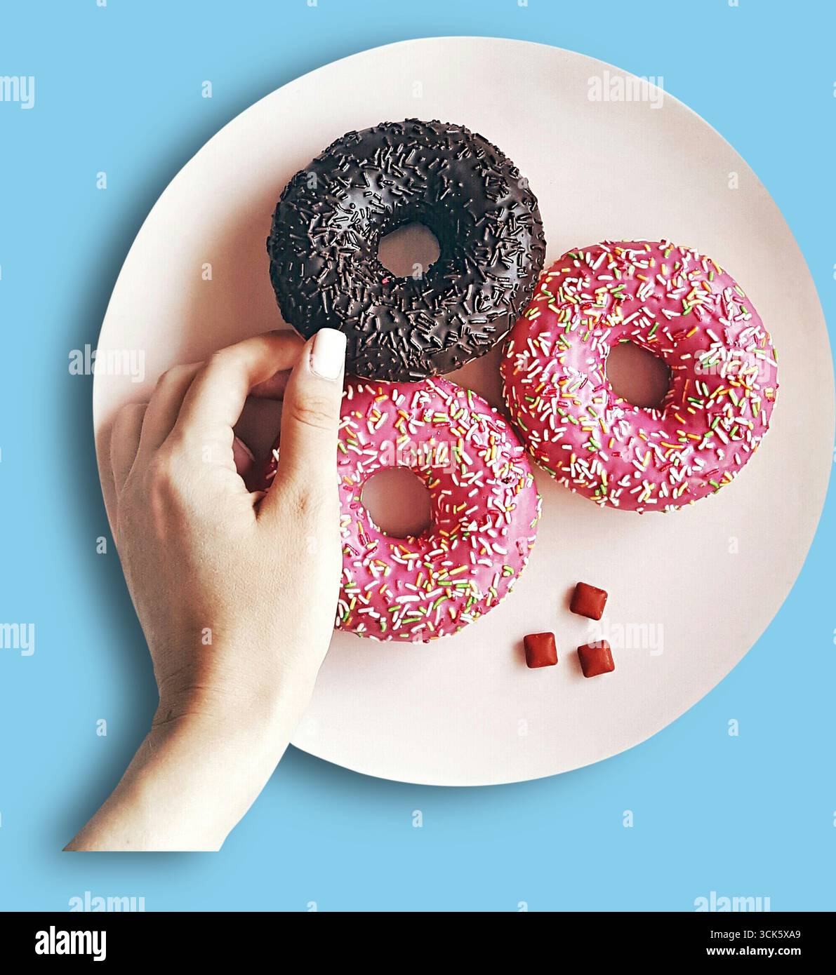 Eine Hand, die auf einem rosafarbenen Teller nach bunten Donuts greift und eine köstliche Auswahl an Leckereien präsentiert. Stockfoto