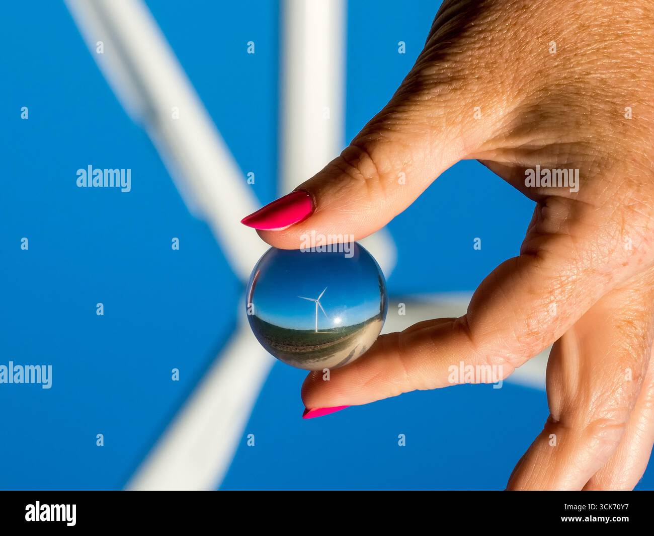 Nahaufnahme einer Hand mit bemalten Nägeln, die eine kleine Glaskugel hält, die eine Windturbine gegen einen klaren blauen Himmel reflektiert Stockfoto