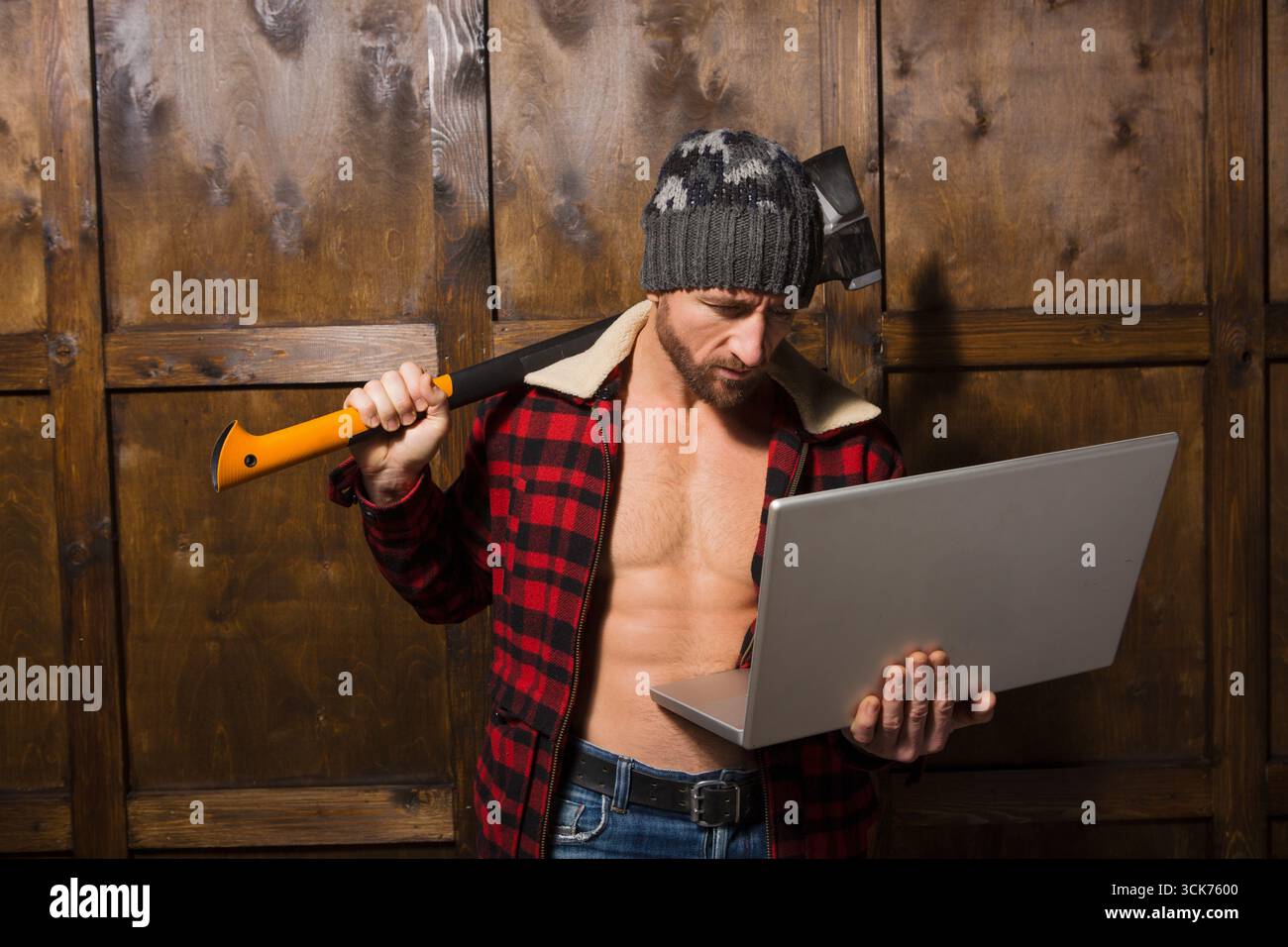 Ein robuster Mann, gefangen zwischen Zuneigung und Frustration, kämpft mit defekten elektronischen Geräten. Er hält eine Axt und einen Laptop, verkörpert den Kampf o Stockfoto