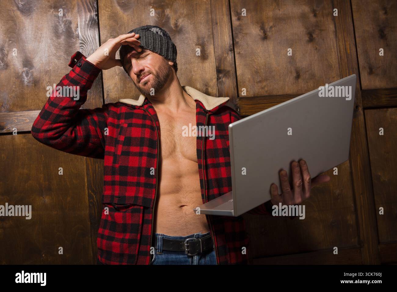 In einer rustikalen Holzkabine greift ein robuster Mann mit einem charmanten, aber frustrierten Ausdruck mit seinem Laptop und zeigt die Mischung aus Romantik und Technol Stockfoto
