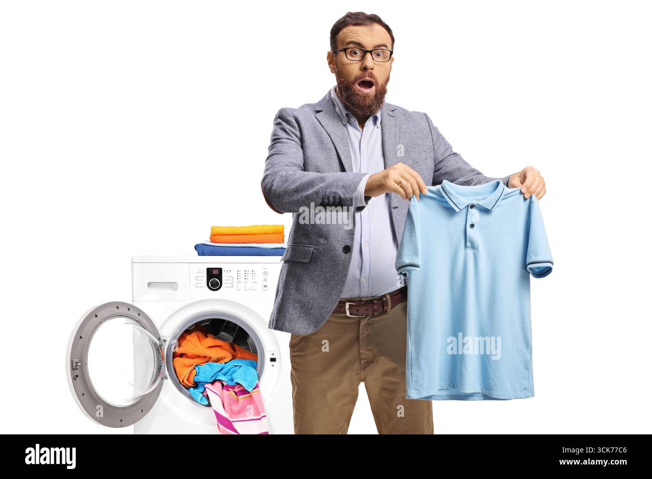 Überraschter Mann, der ein kleines T-Shirt vor einer Waschmaschine auf weißem Rücken hält Stockfoto