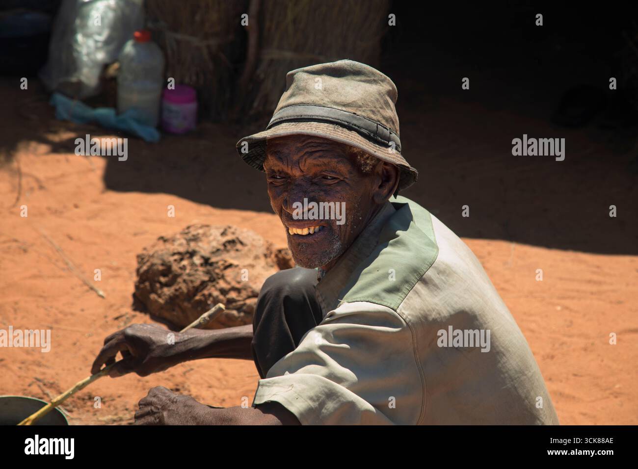 Afrikanischer alter Mann mit Hut, der auf offenem Feuer Essen kocht, sitzt vor seiner Strohhütte in Botswana, Flüchtlingslager Stockfoto