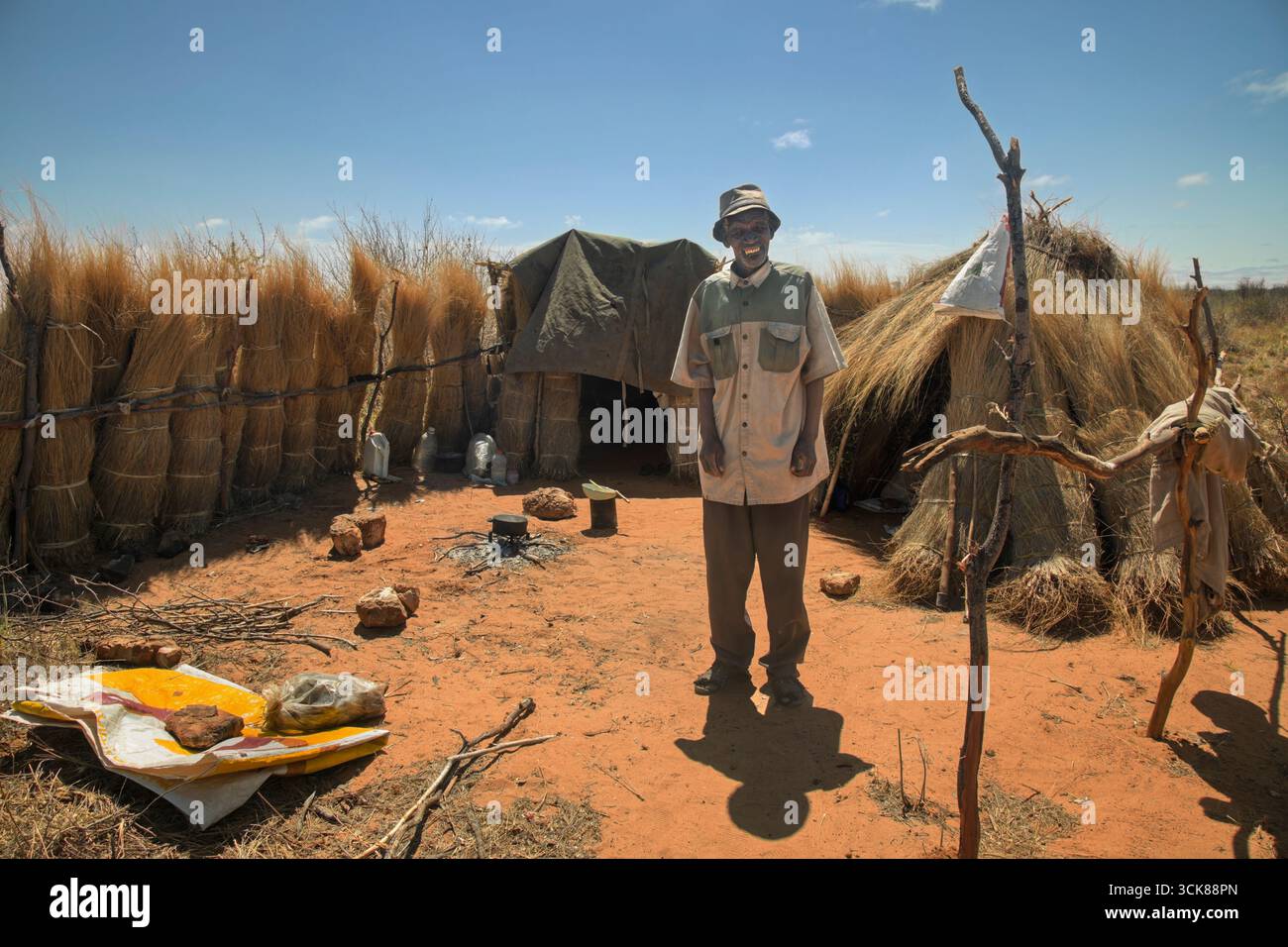 Afrikanischer alter Mann mit Hut, der auf offenem Feuer Essen kocht, sitzt vor seiner Strohhütte in Botswana, Flüchtlingslager Stockfoto