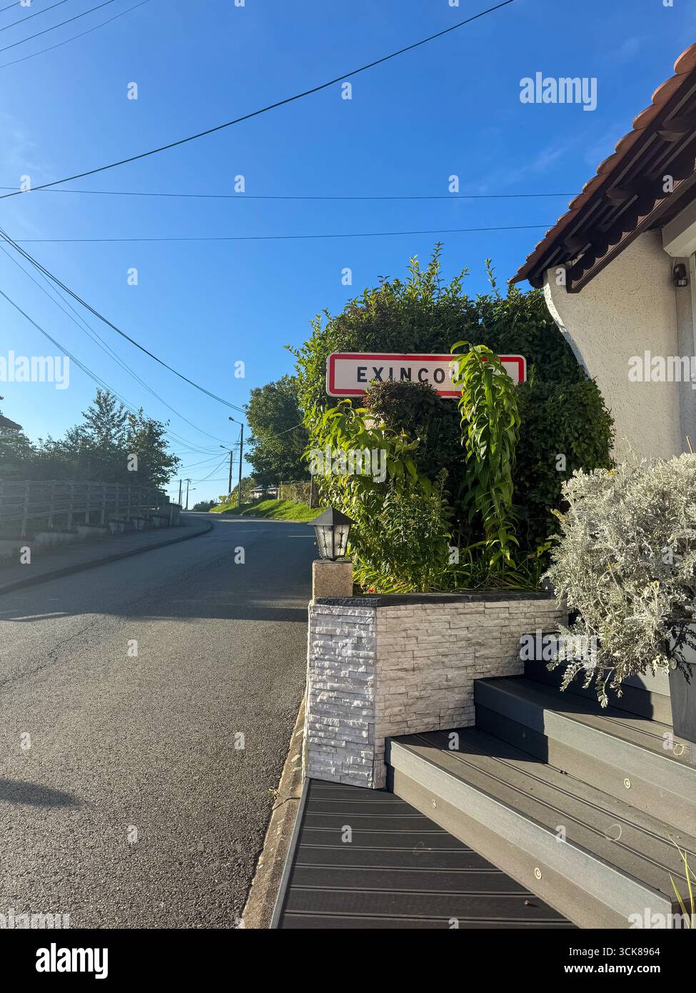 Ruhige Straße in Exincourt, Frankreich, mit einer geschwungenen Straße, Steinmauer, Stadtschild, charmanten Häusern, kleinen Gärten, üppiges Grün und sonniger blauer Himmel - Smartphone-aufgenommenes Stockfoto