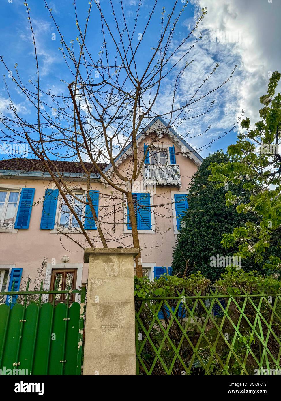 Bezauberndes französisches Haus mit hellblauen Fensterläden, verziertem Balkon und Gartenbäumen unter einem teilweise bewölkten Frühlingshimmel. - Smartphone-aufgenommenes Stockfoto