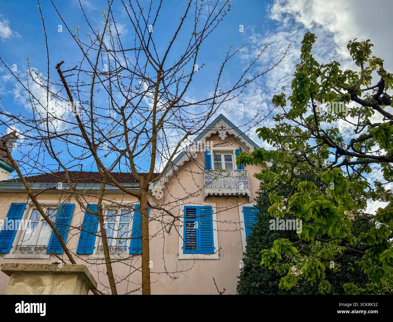 Bezauberndes französisches Haus mit hellblauen Fensterläden, verziertem Balkon und Gartenbäumen unter einem teilweise bewölkten Frühlingshimmel. - Smartphone-aufgenommenes Stockfoto