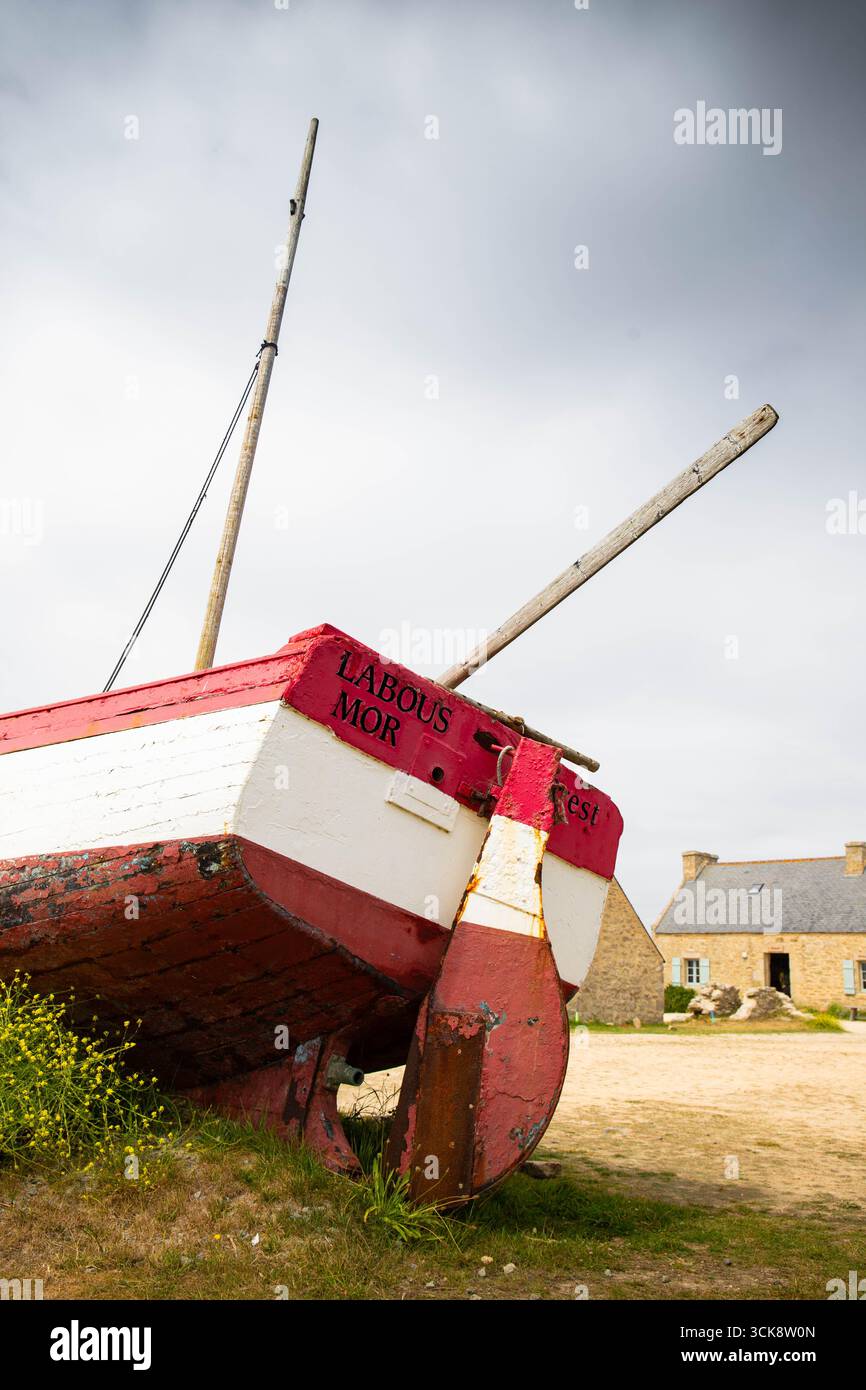 Rotes verlassenes Fischboot im alten Fischerdorf Meneham, Bretagne, Frankreich, mit strohgedeckten Hütten im Hintergrund. Ruderansicht mit Wolken, n Stockfoto