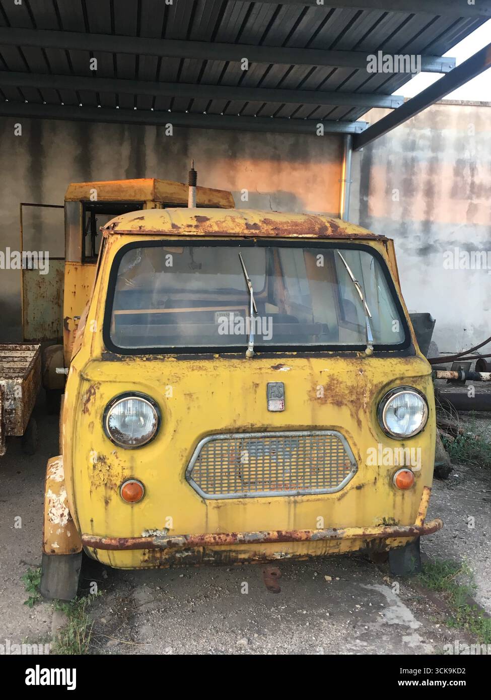 Italien – 30. August 2025: Ein rostiger Oldtimer wurde auf einem ländlichen italienischen Hof aufgegeben, mit verwitterter Karosserie, Platten Reifen und bewachsenem Unkraut unter klarem Licht Stockfoto