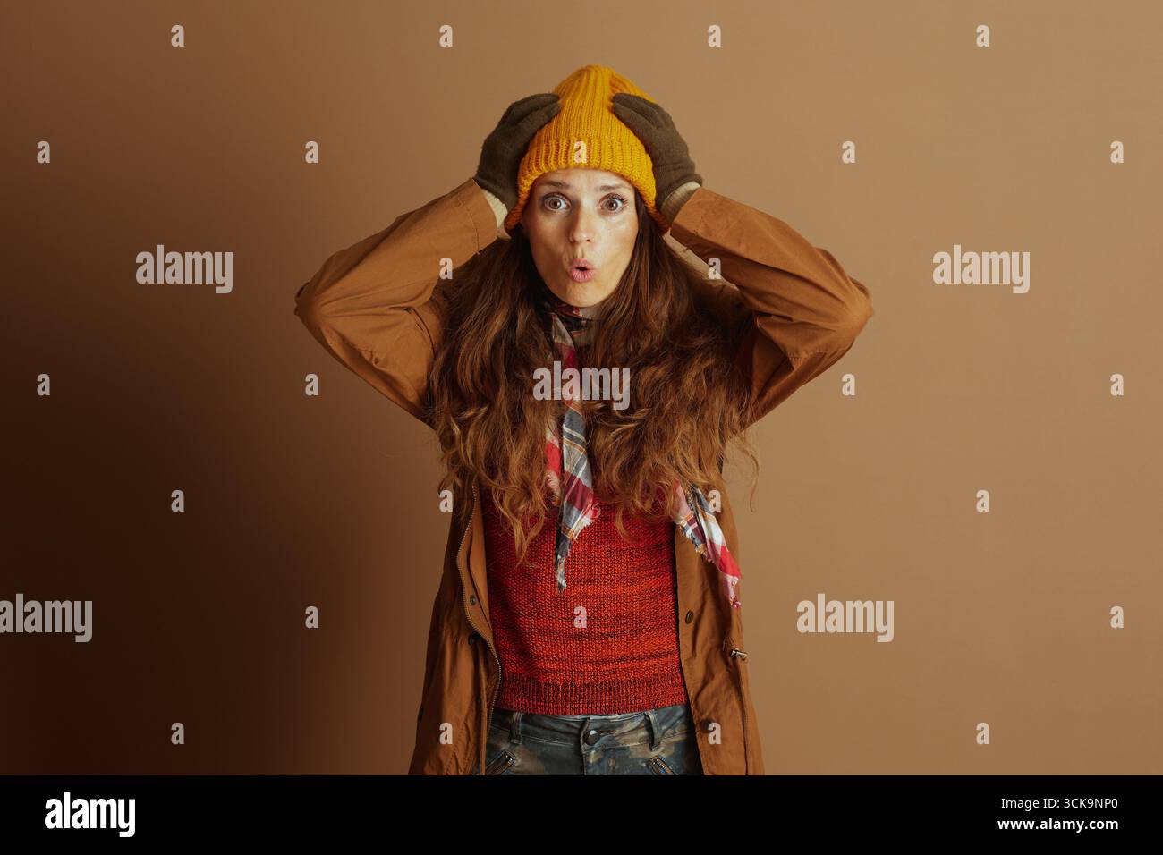 Eine Frau in kuscheliger Herbst-Winter-Kleidung, einschließlich Trenchcoat und Mütze, schaut den Betrachter mit einem breiten, überraschten Ausdruck, der den Betrachter direkt ansieht Stockfoto