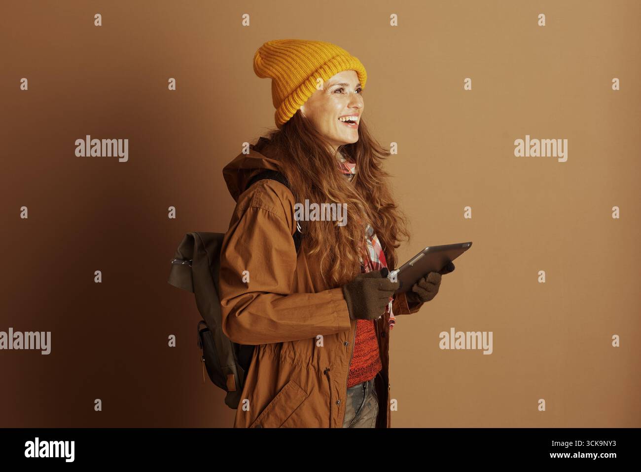 Eine lächelnde Frau mit langen, welligen braunen Haaren, die in einer gelben Mütze und einer braunen Jacke für Reisen gekleidet ist, hält eine Tablette und trägt einen Rucksack, was symbolisiert Stockfoto