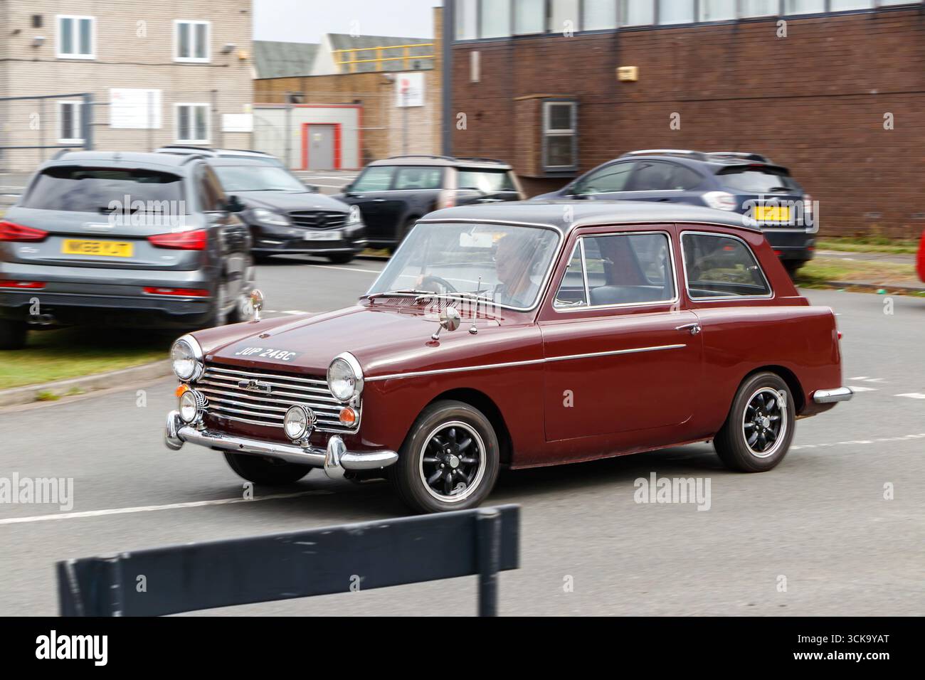 Ein braunes Oldtimer austin A40 fährt auf einer Straße mit modernen Fahrzeugen, die im Hintergrund geparkt sind und eine Mischung aus klassischem und zeitgenössischem Stil bietet Stockfoto