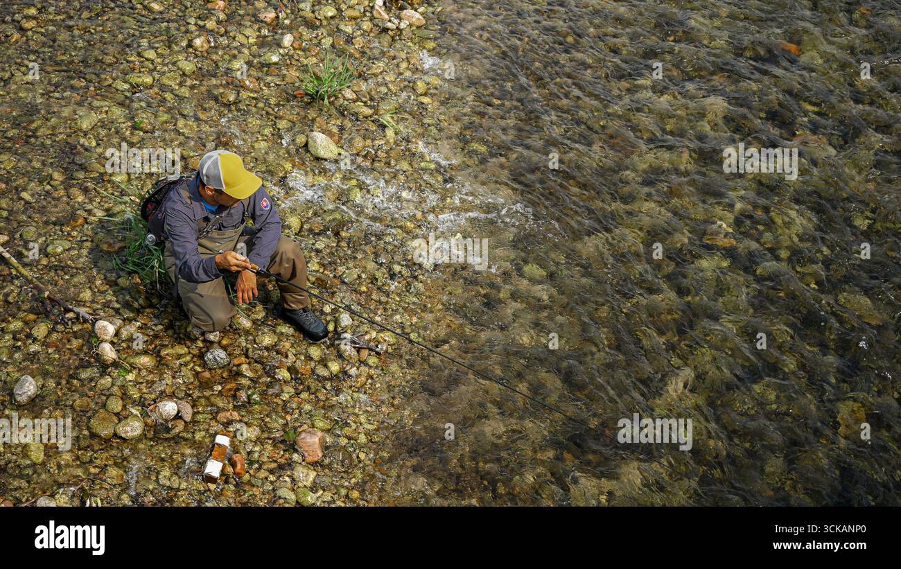 Fischer mit Wathose und Baseballmütze, die auf einem felsigen Flussufer sitzen und Angelschnur in klares Wasser werfen. Stockfoto