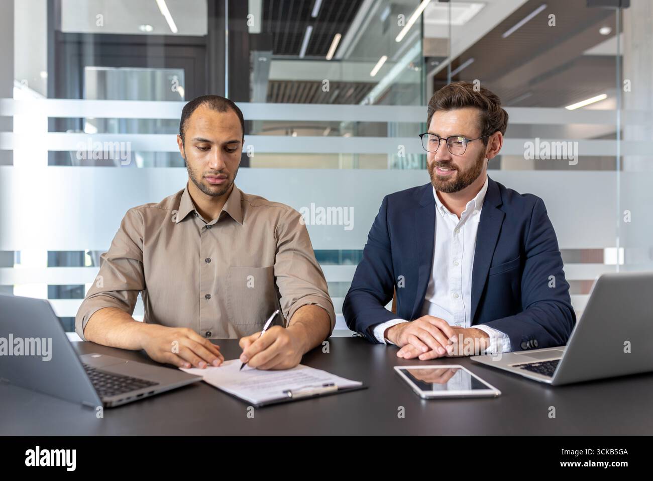 Ein Geschäftsmann prüft und unterzeichnet einen Vertrag sorgfältig, während sein Vorgesetzter in einer modernen Büroumgebung beobachtet und Ratgeber gibt, indem er Laptops und Tablets für einen effizienten Workflow nutzt Stockfoto
