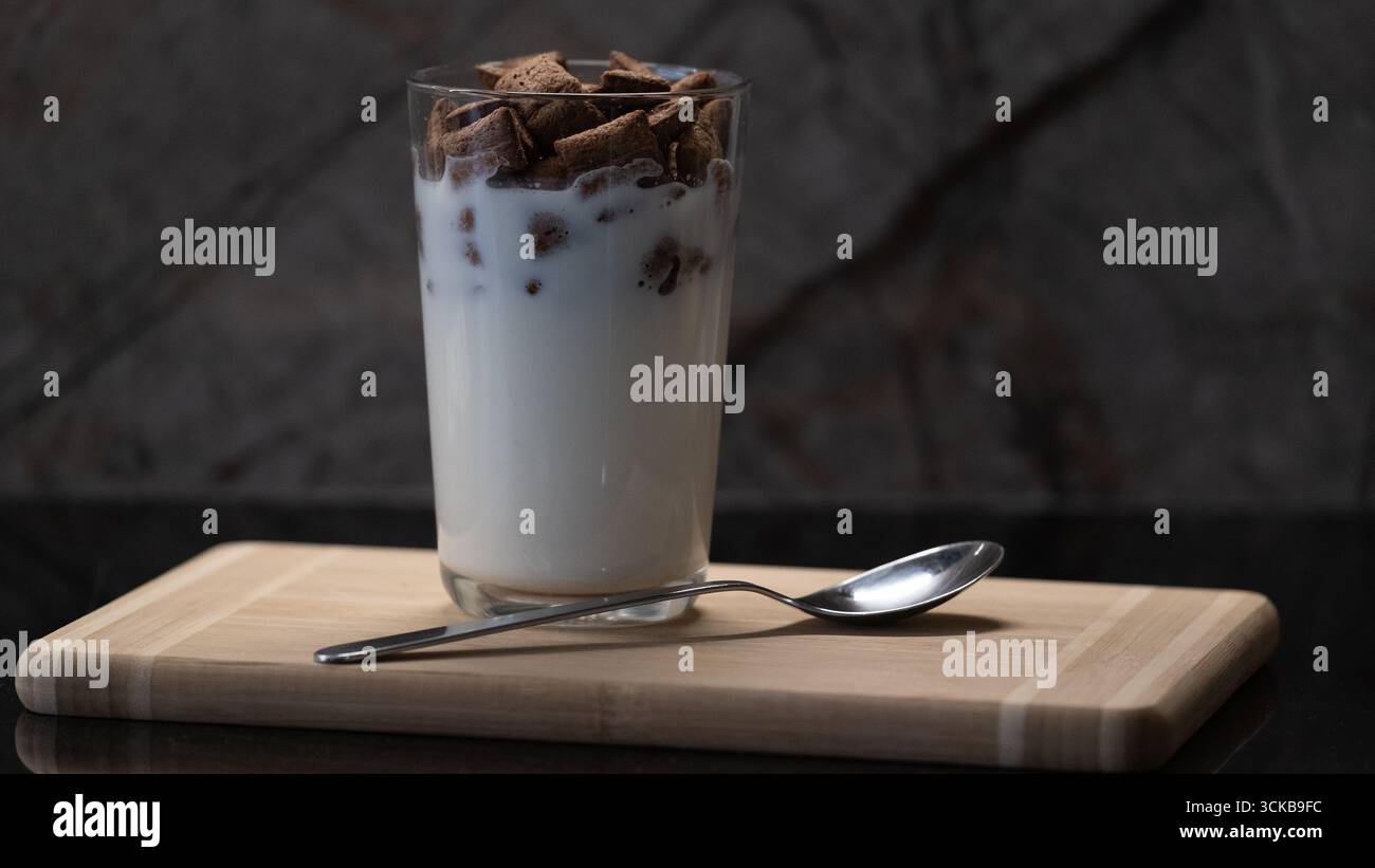 Nahaufnahme eines Glases, das mit Milch und Schokoladen-Müslistücken gefüllt ist, auf ein hölzernes Schneidebrett mit einem Metalllöffel gelegt. Die Komposition ist hochgradig Stockfoto