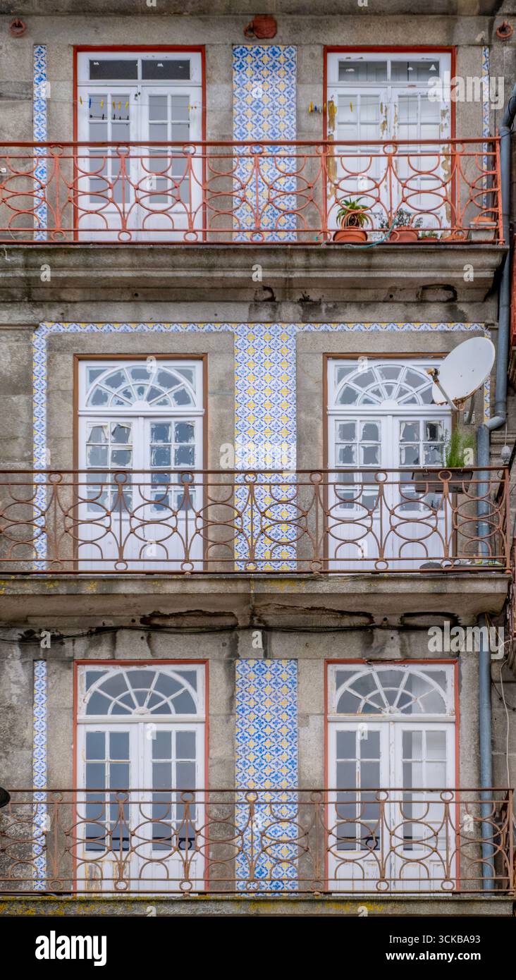 Wohnfassade mit Bogenfenstern, vertikalen Azulejo-Streifen und verzierten Balkonen in Porto, Portugal. Blaue und gelbe Fliesen, Satellitenschüssel Stockfoto